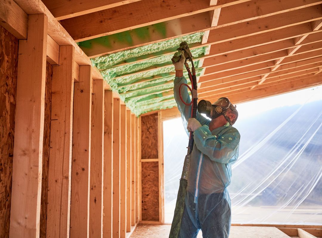 Man spraying green foam insulation on ceiling joists in a building under construction.