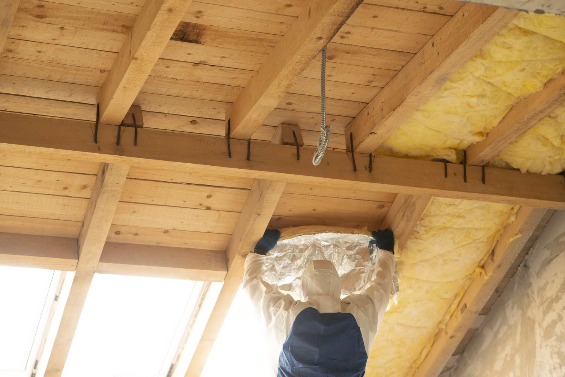 Person installing insulation in an attic with wooden beams, wearing protective gear. Yellow insulation visible.
