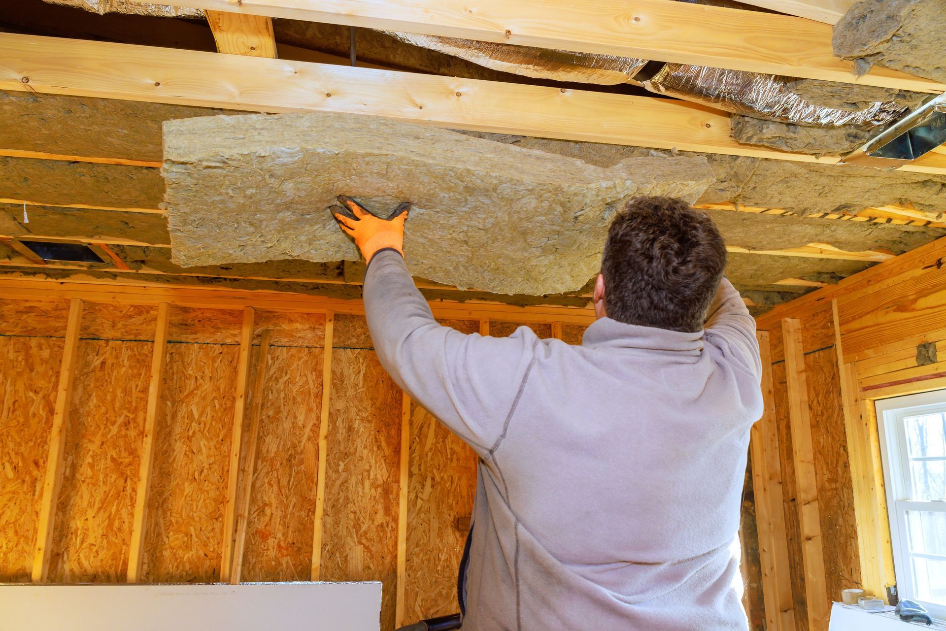 Person installing insulation in a wooden framed ceiling; wearing gloves and a grey sweatshirt.