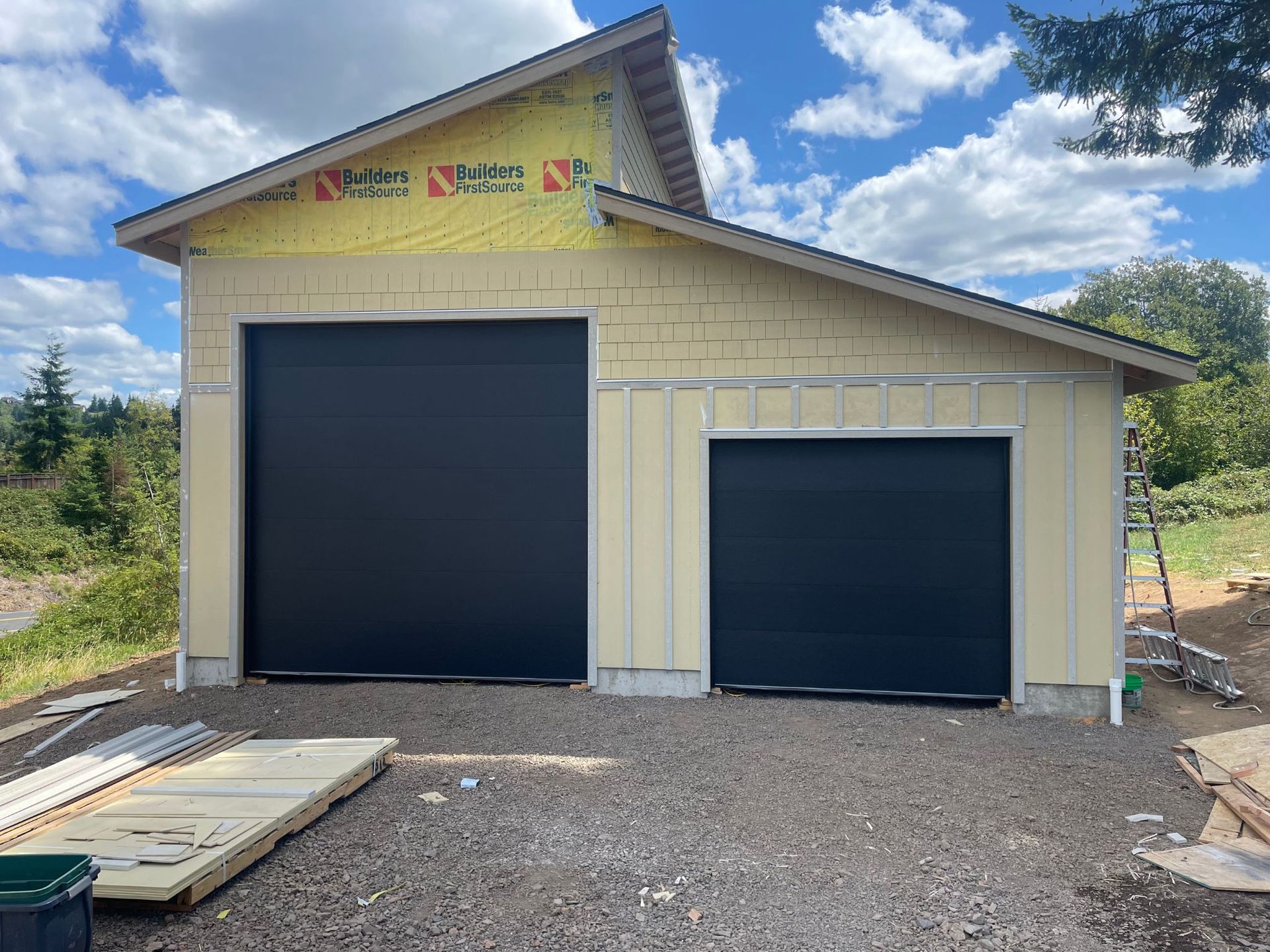 New building with two black garage doors, yellow siding, gravel ground, and construction materials.