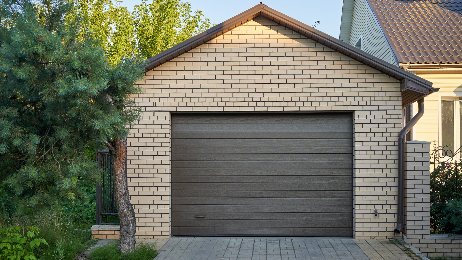 A brick garage with a brown garage door is next to a house.