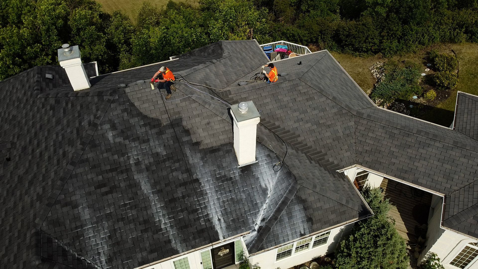 Two workers in orange suits on a dark shingled roof, applying a white substance. Green trees and sky in background.