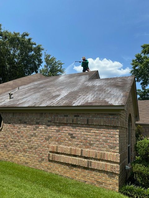 Person pressure washing a roof; brown brick house, blue sky.