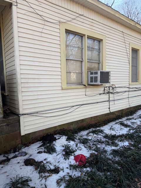 A light yellow house with two windows and an AC unit. The ground is covered in snow.