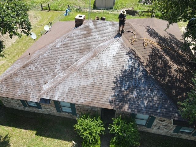 A person power washing a shingle roof, removing dark streaks. Green grass surrounds the house.