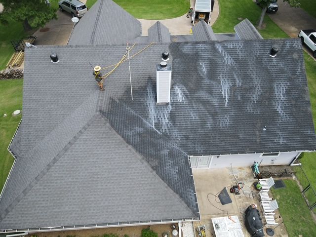 Person on a roof washing it with a hose. Gray roof, white residue, green lawn.