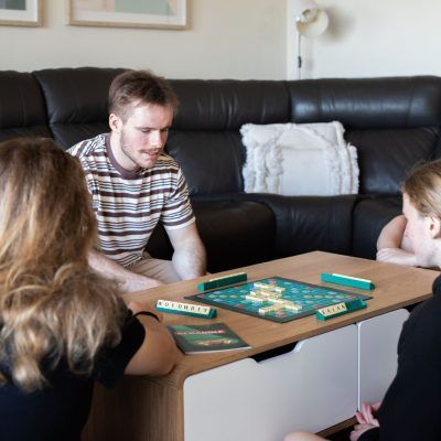 Three people playing a board game around a coffee table in a living room. One man and two women are focused on the game.