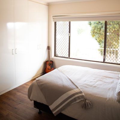 Bedroom with a bed, guitar, window, and white wardrobe. The bed has a gray blanket, and the room has wooden floors.