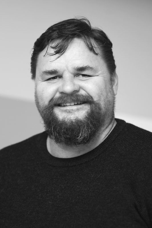 Man with a full beard smiles, wearing a dark sweater. He is indoors.