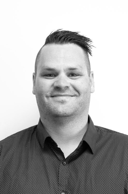 A man with a mohawk hairstyle and a dark collared shirt smiles at the camera against a white background.