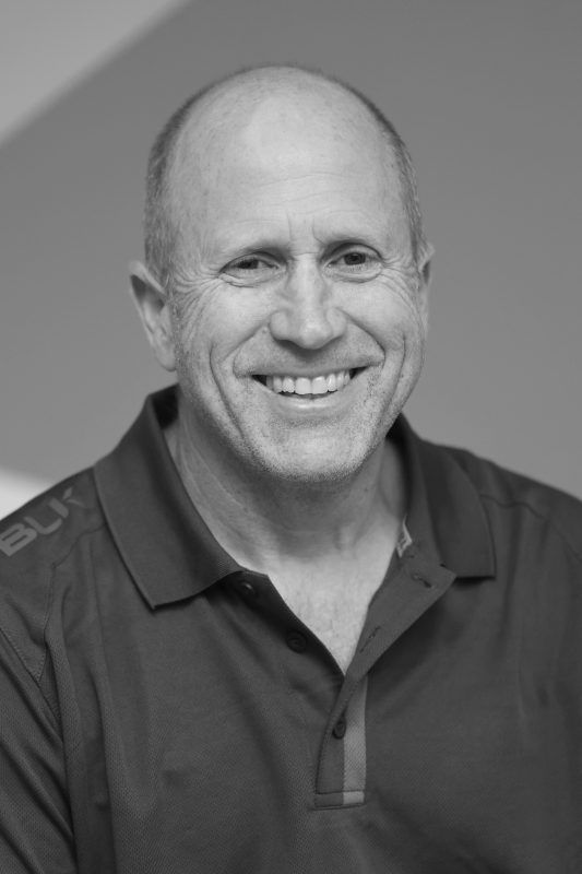 Smiling, balding man in a polo shirt, photographed from the chest up against a blurred background.
