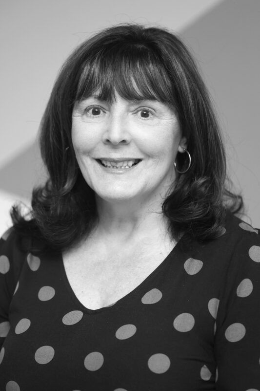 Woman with dark hair and a polka-dot top smiles at the camera. She wears hoop earrings and has a plain background.