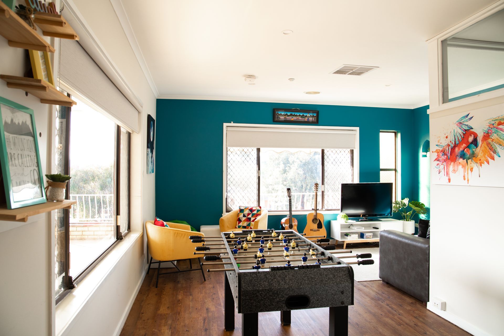A room with a foosball table, TV, teal accent wall, and large windows overlooking trees. Two guitars lean against the wall.