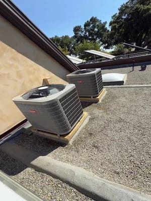 Two outdoor air conditioning units on a gravel rooftop beside a beige wall