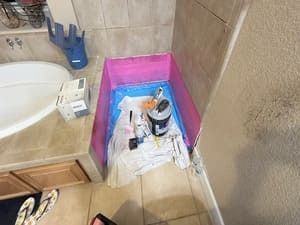Bathroom corner with a pink-lined shower area under repair, cleaning supplies, and tools on the floor