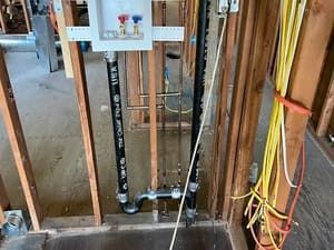 Exposed plumbing and electrical wiring in a framed construction wall with a white utility panel.