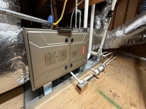 Beige furnace and ductwork in an attic crawlspace with insulated silver vents and exposed pipes.