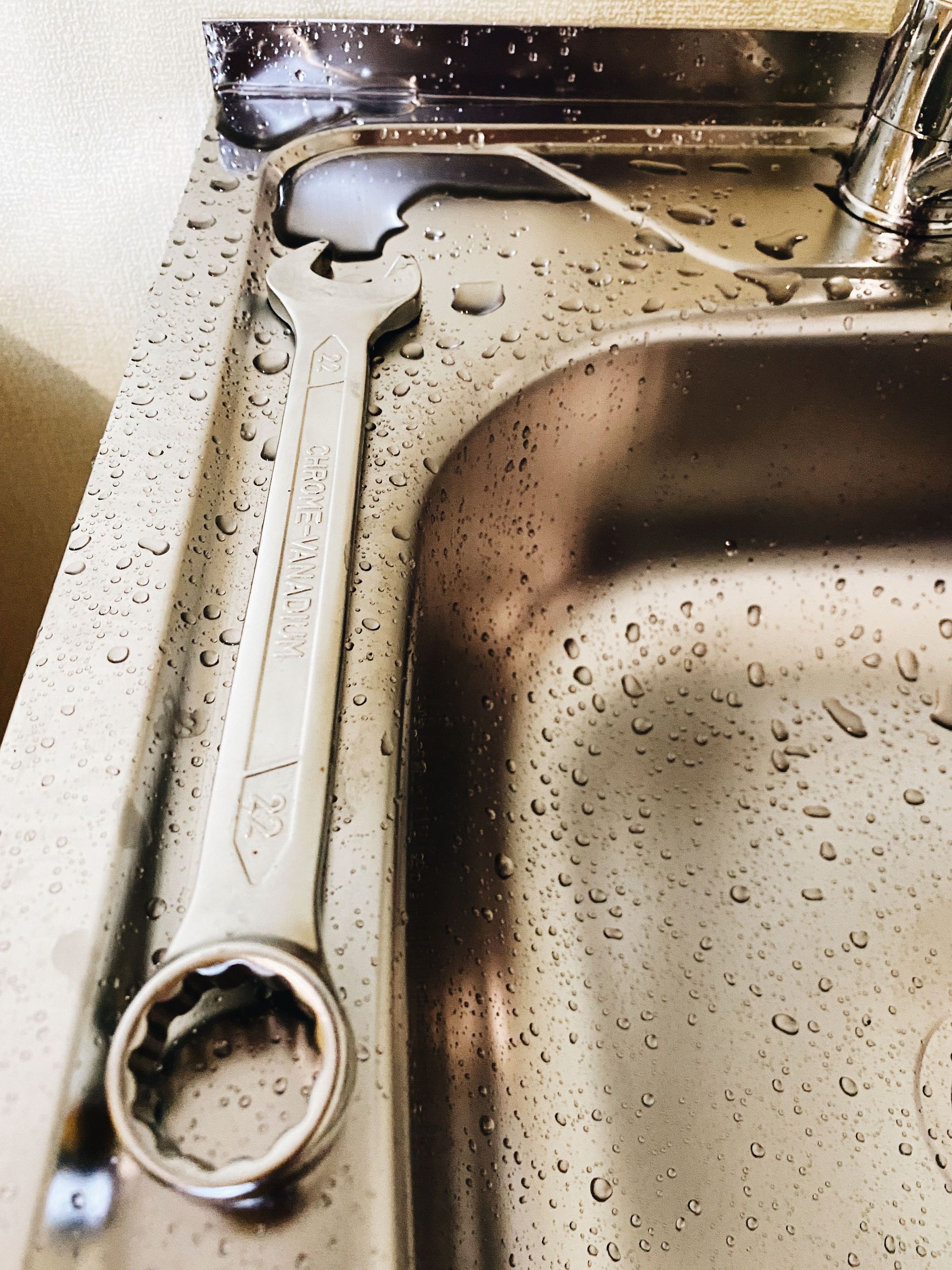 Large wrench resting beside a wet stainless-steel sink with water droplets and a faucet nearby