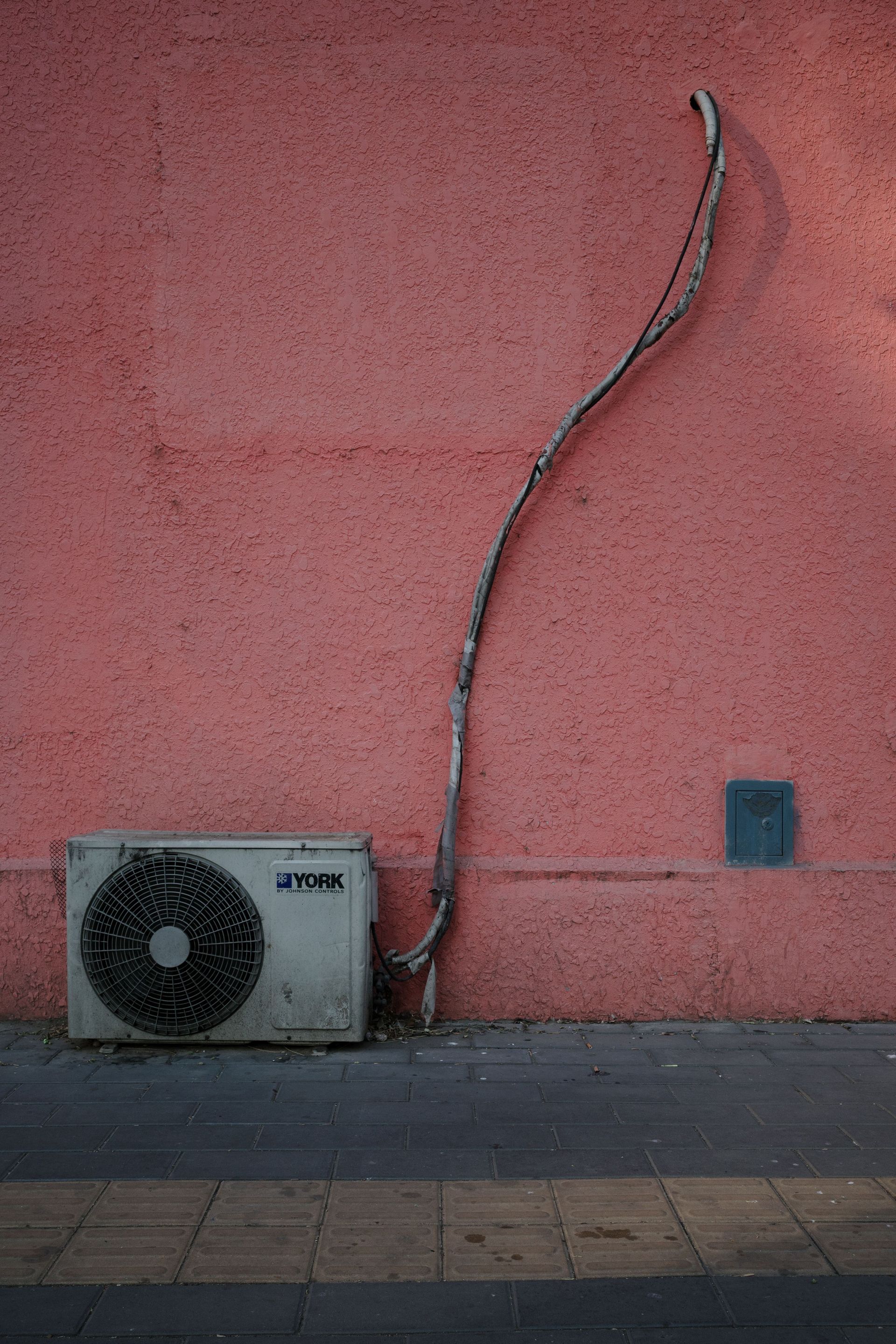 Outdoor wall with a wall-mounted air conditioner and exposed conduit on a pink stucco facade.
