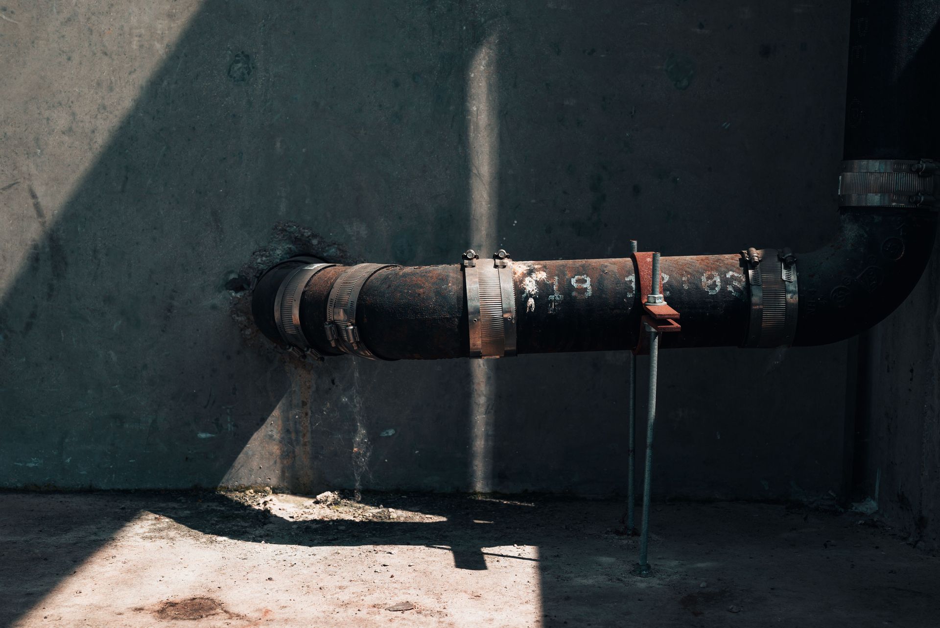 Rusty black pipe running through a gray concrete wall with sunlight casting shadows.