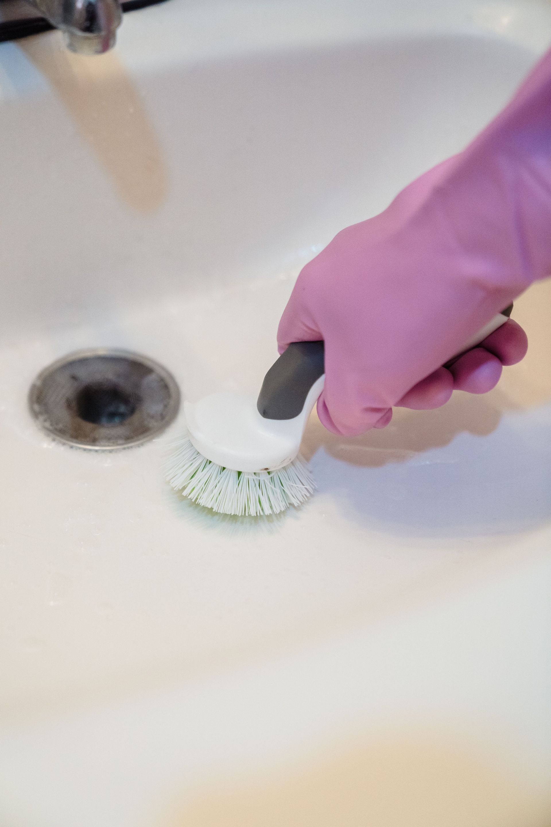 Pink-gloved hand scrubbing a white sink with a brush near the drain