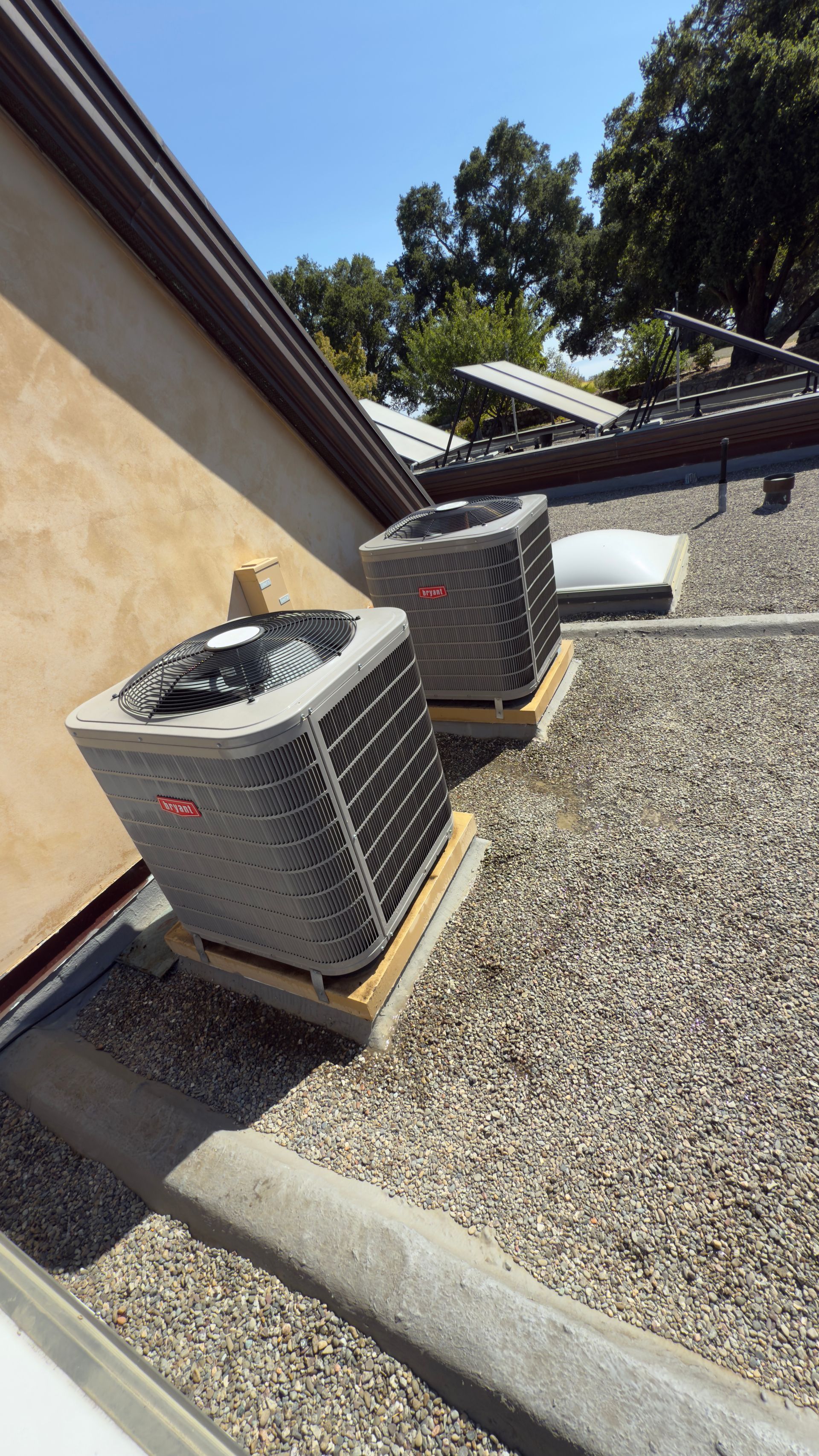 Two outdoor HVAC units on a gravel rooftop beside a beige wall and trees