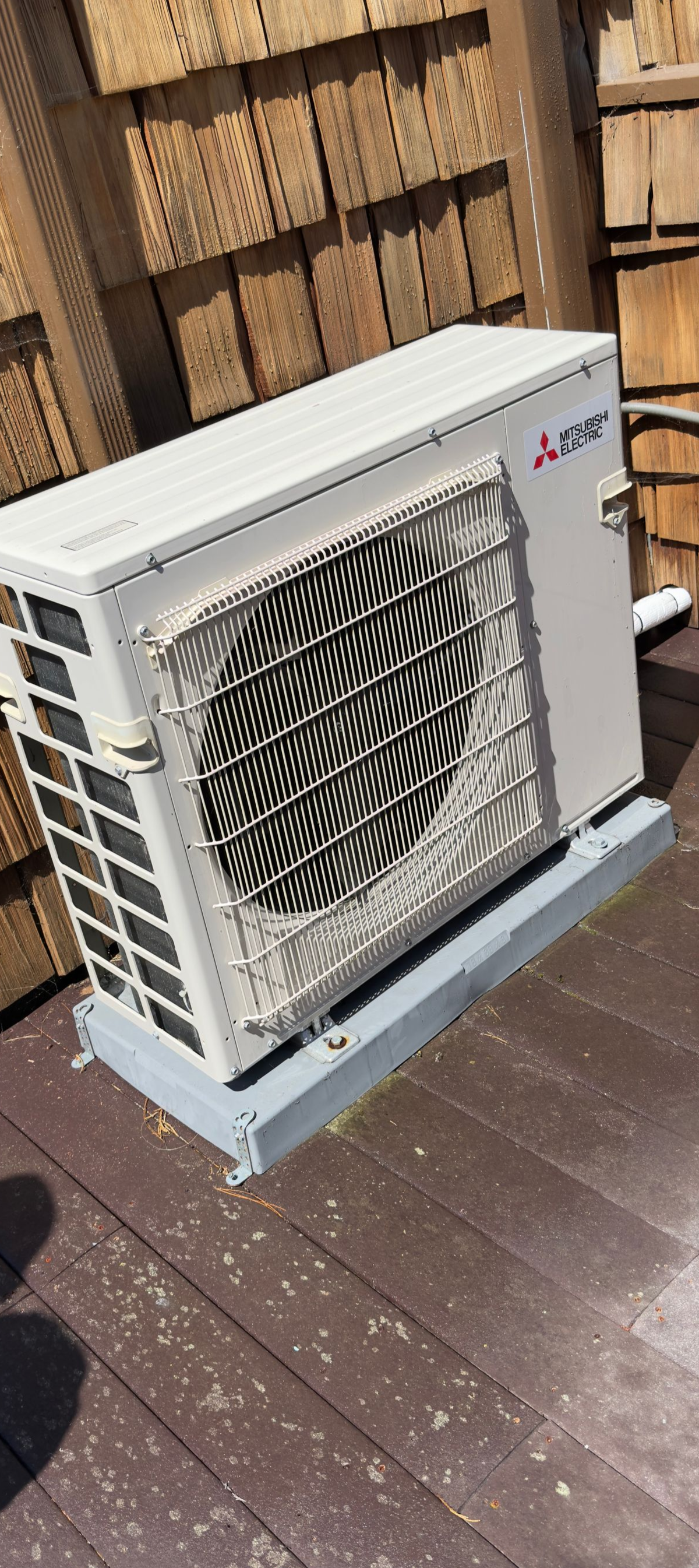 Outdoor heat pump unit beside a wooden fence, with a large fan grille and white casing.