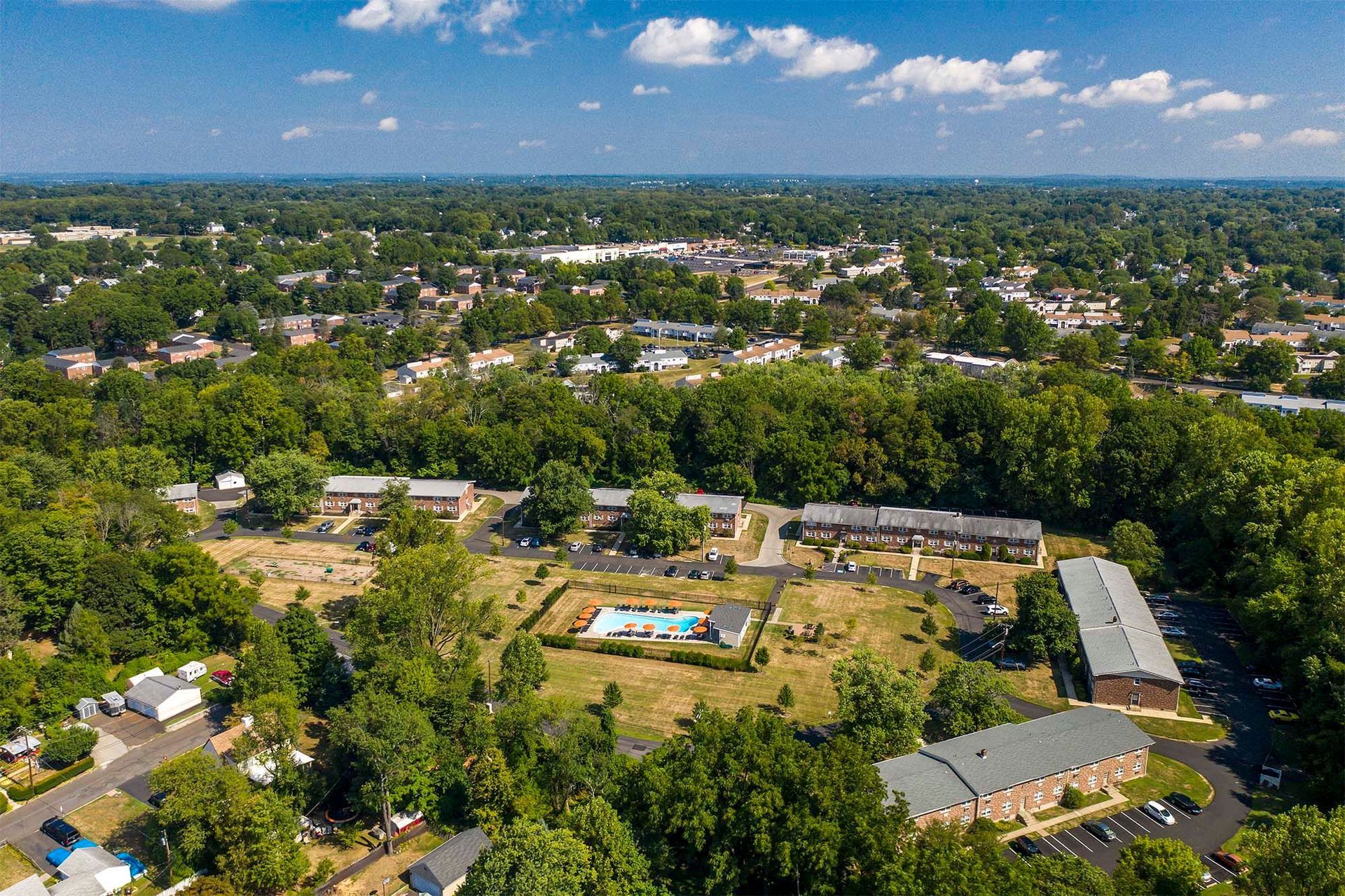 Aerial view of a suburban apartment community with brick buildings, a central pool, and plenty of trees.