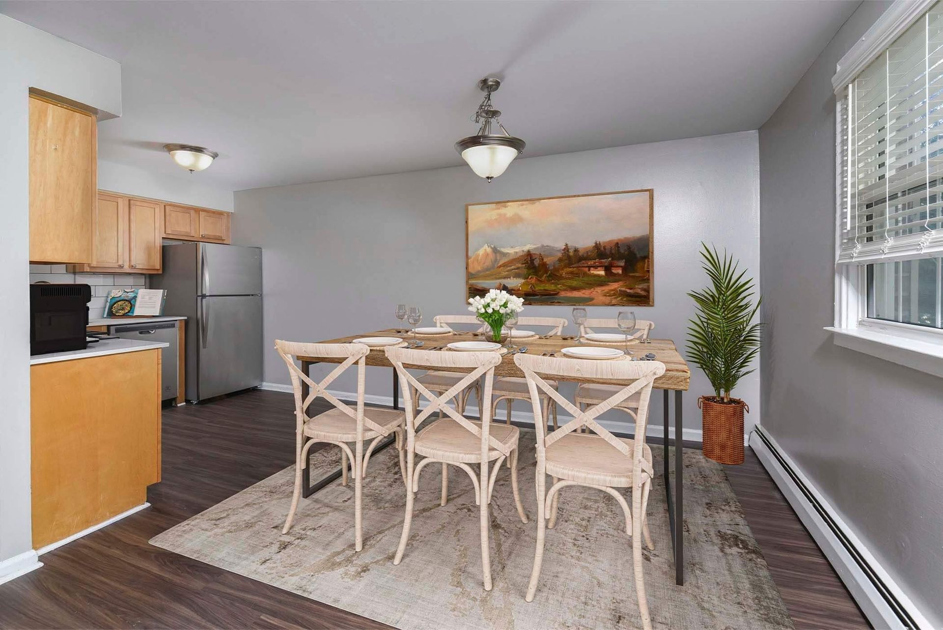 Dining area in an apartment with a wooden table, six chairs, a wall painting, and a plant by the window.