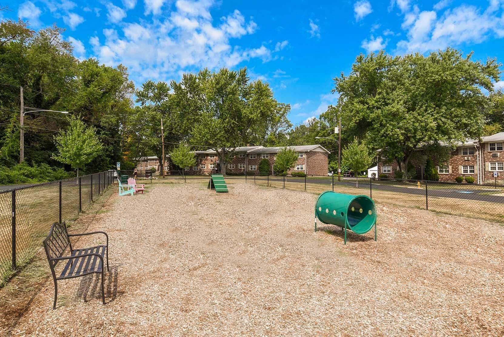 Fenced playground area with benches and green play equipment near apartment buildings.