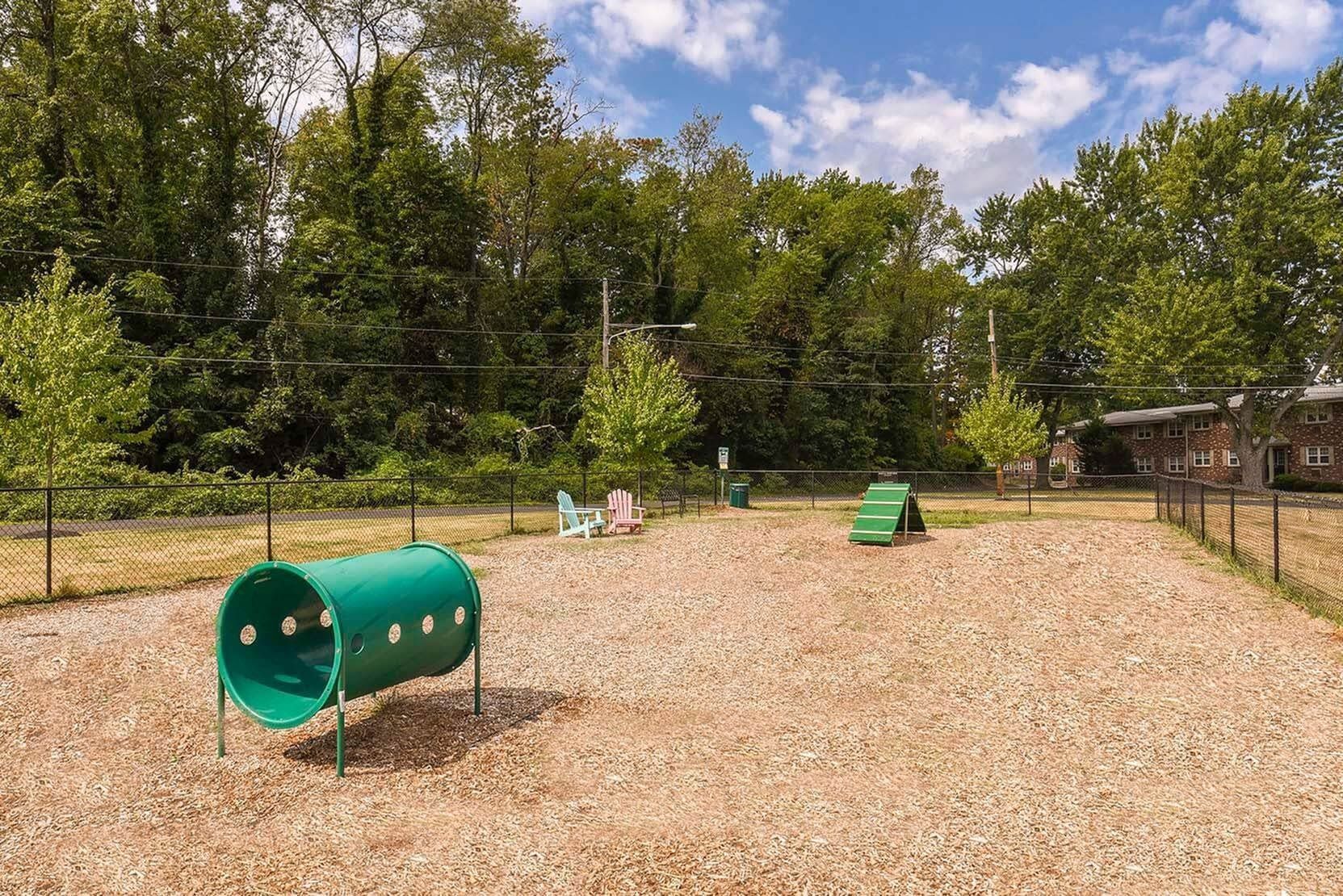 Playground area with a green tunnel, a slide, and seating behind a chain-link fence.