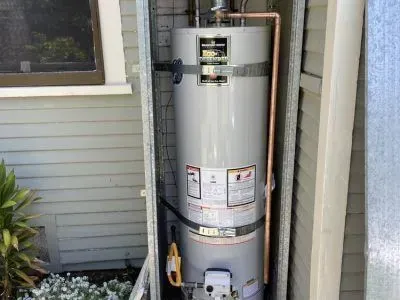 A water heater is sitting outside of a house next to a window.