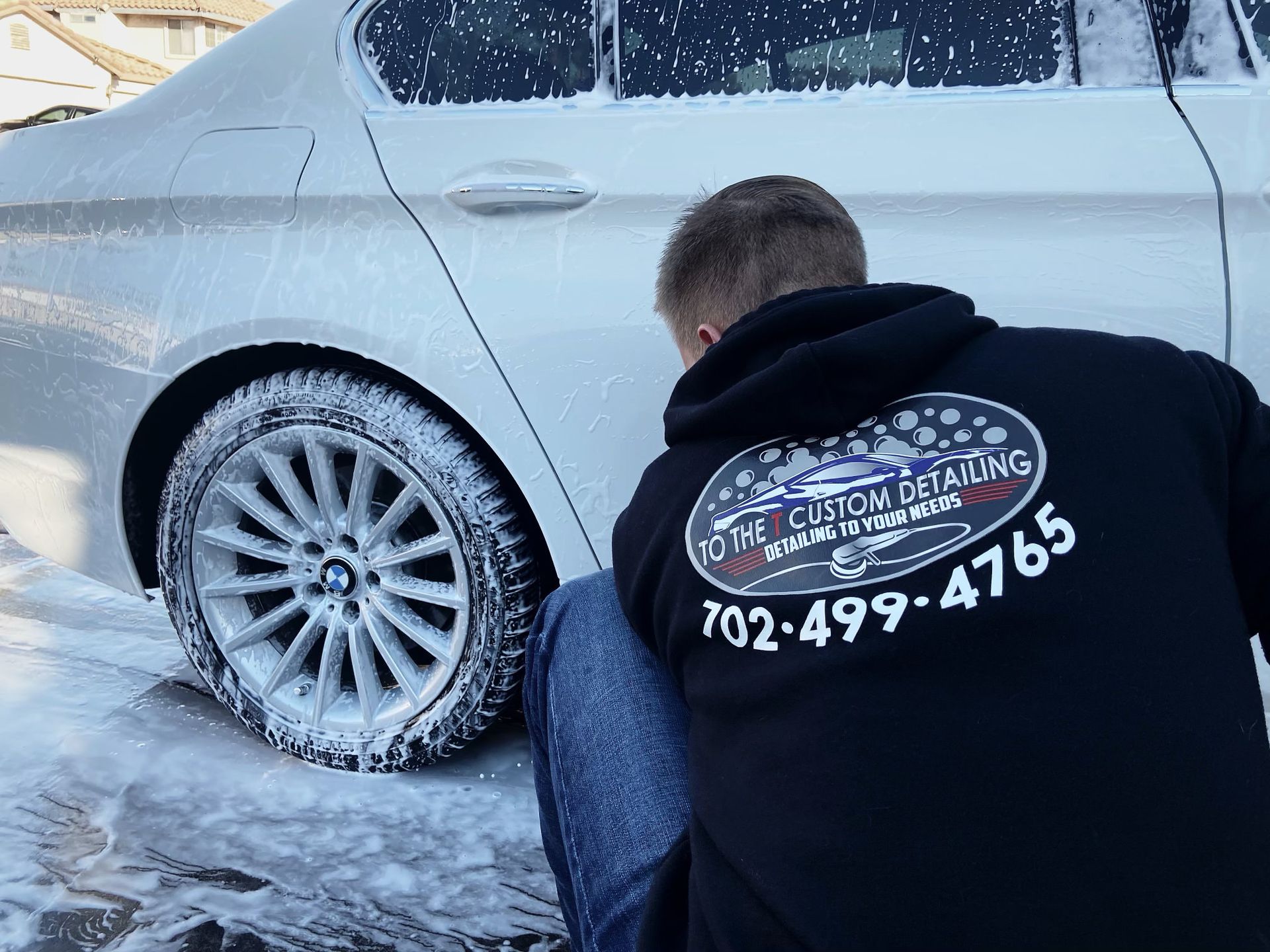 A man is kneeling down next to a white car covered in foam.