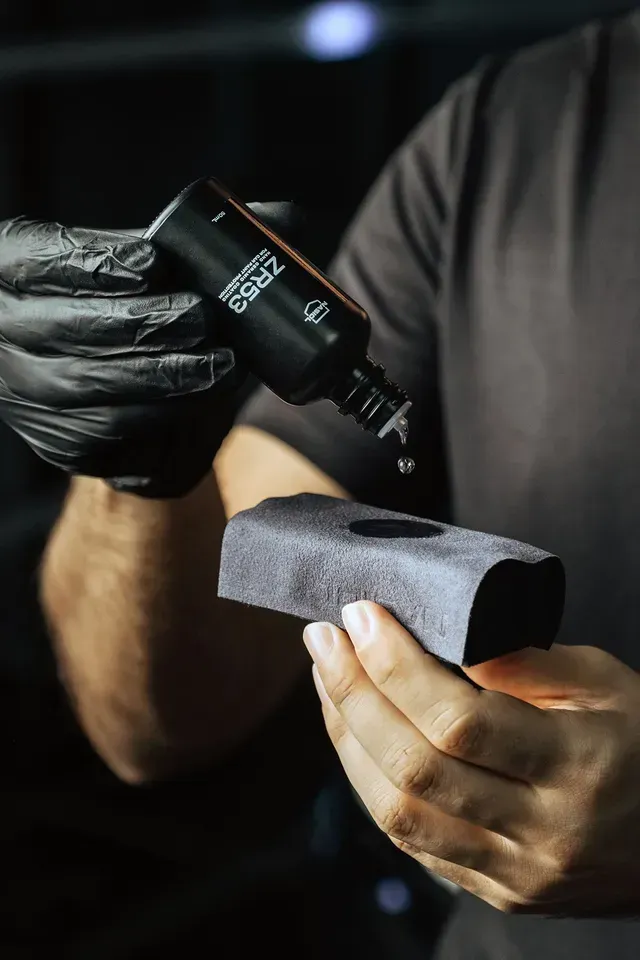 A person in black gloves applies a liquid ceramic coating from a bottle onto a foam applicator pad.