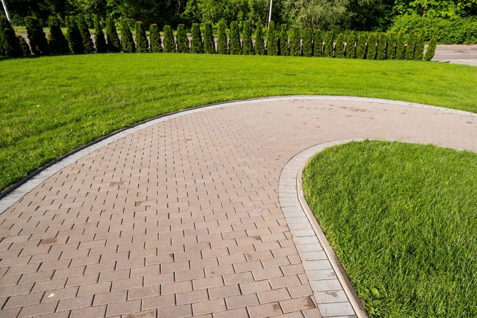 A paved stone walkway curves through a lush green lawn, bordered by a row of evergreen shrubs in the background.