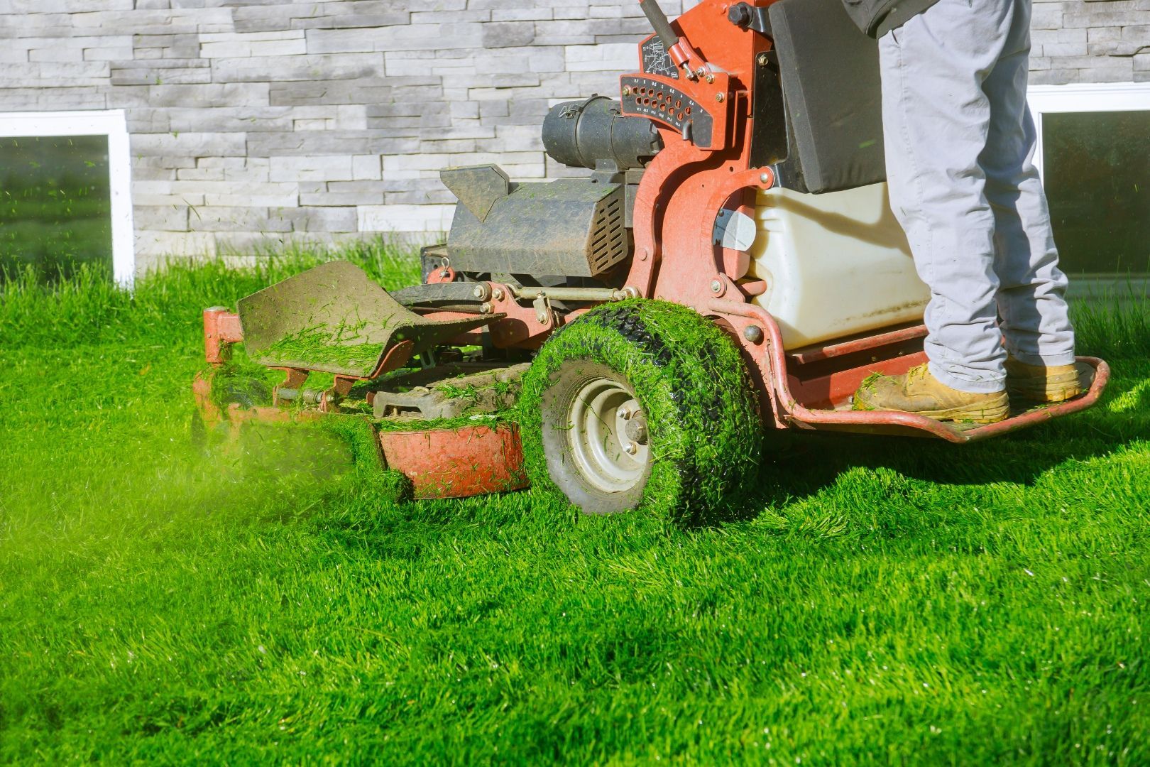 A person operates an orange stand-on commercial lawn mower on a green lawn in front of a house with stone siding.