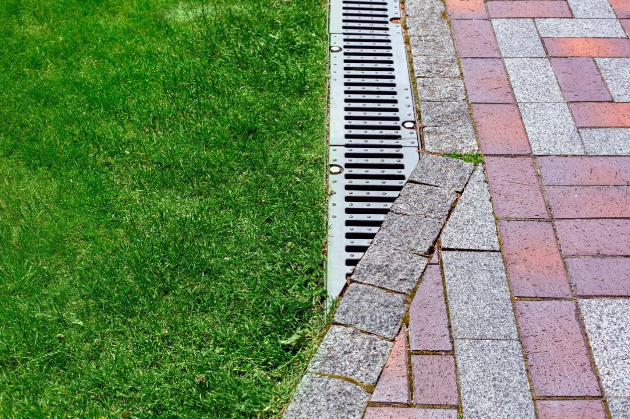 A metal trench drain separates a green grass lawn from a paved walkway made of red and grey bricks.