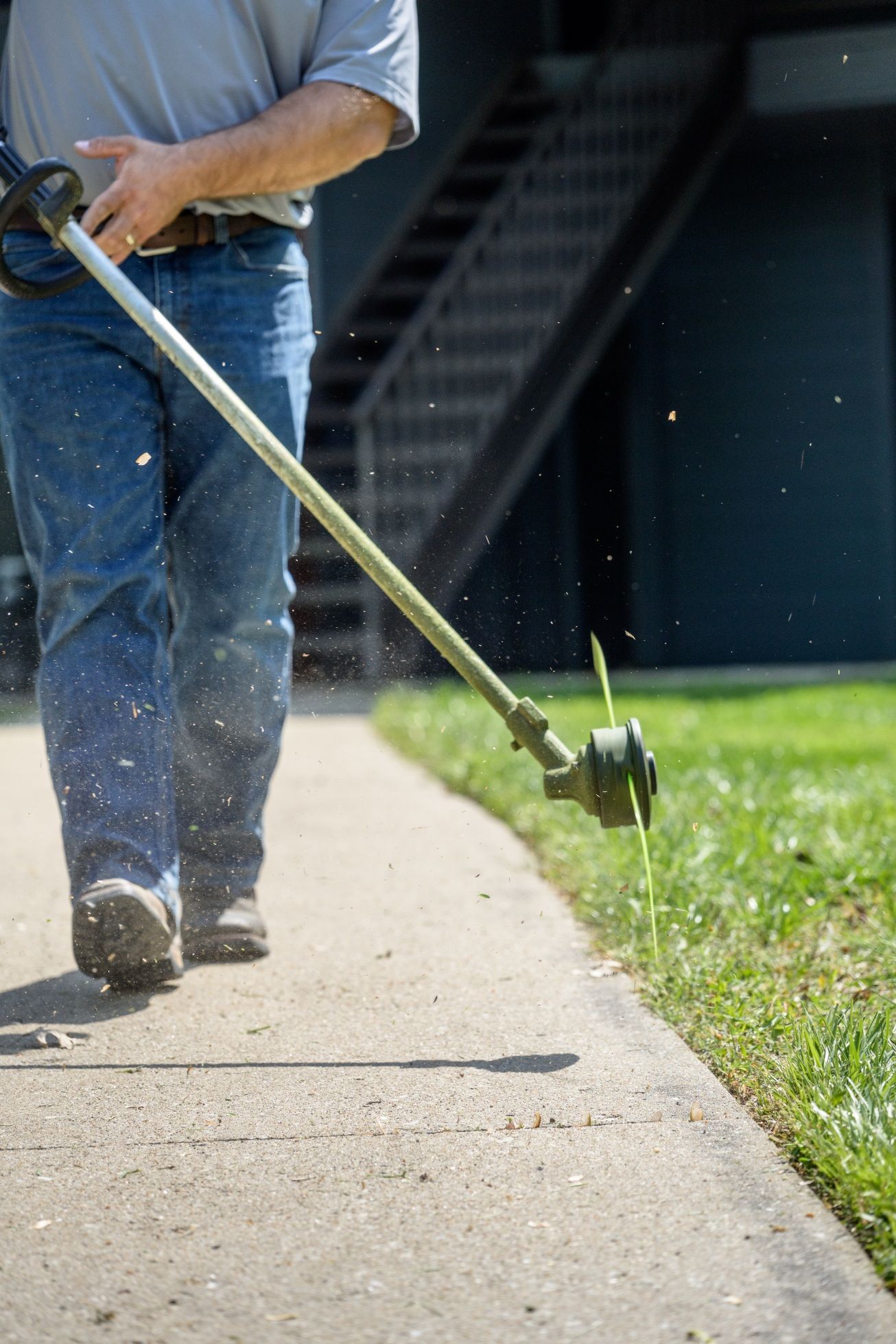 A person in jeans and a gray shirt uses a string trimmer to edge a green lawn along a concrete sidewalk.
