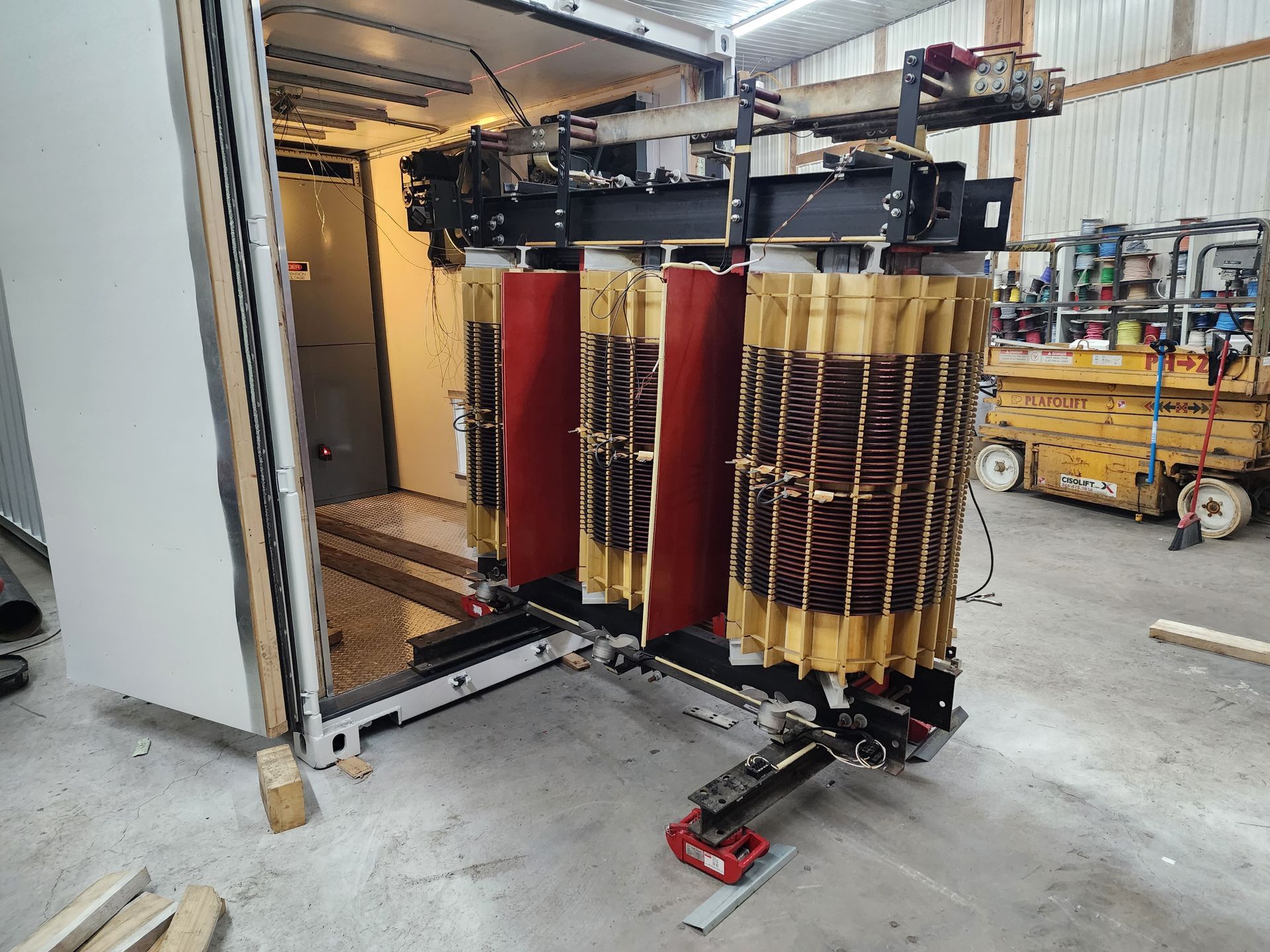 Un gros noyau et des bobines de transformateur industriel triphasé, partiellement sortis d'un conteneur maritime sur des rails métalliques.