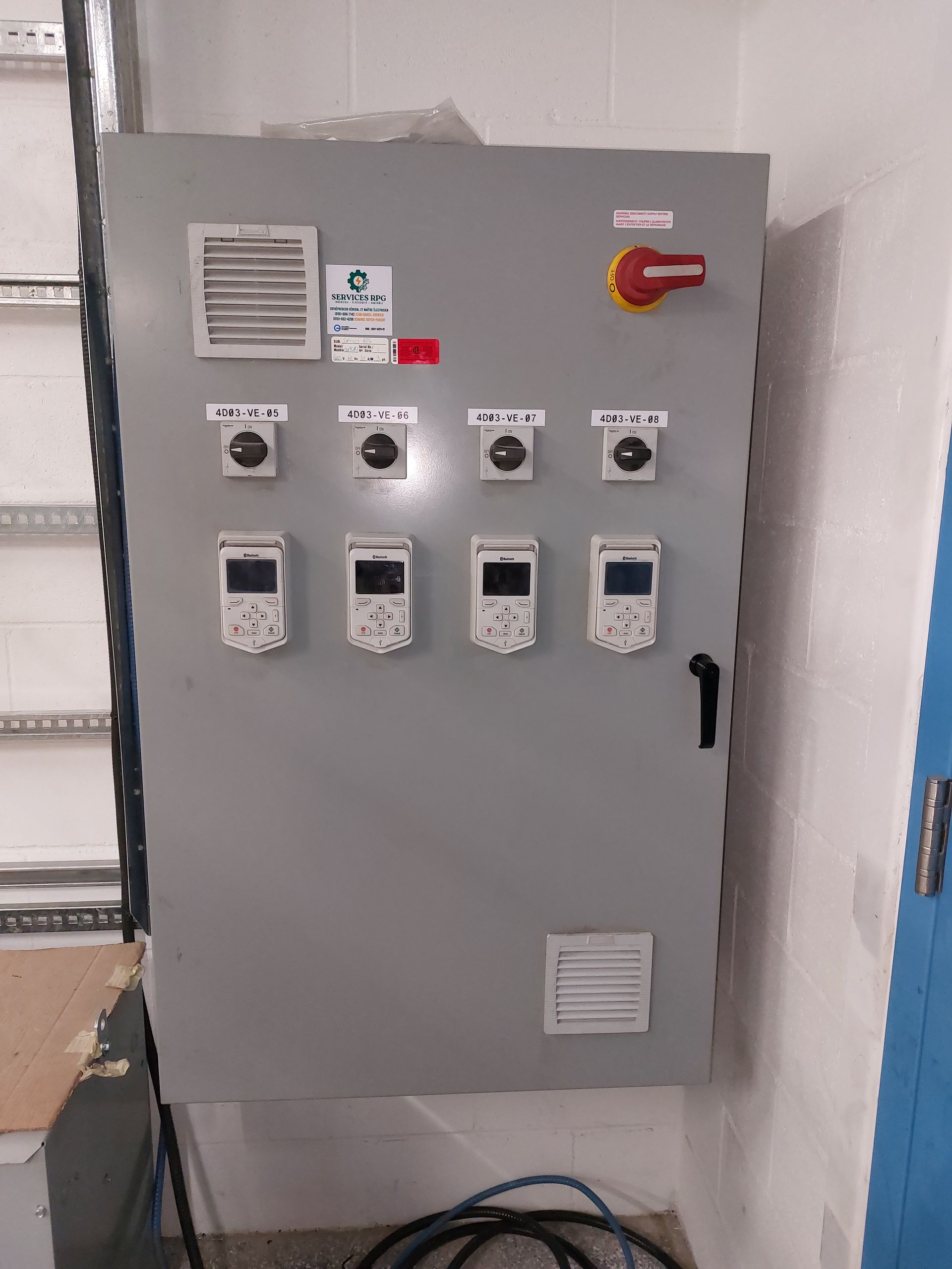 Une armoire électrique industrielle grise, montée sur un mur blanc, comportant quatre interrupteurs, des affichages numériques et des aérations.