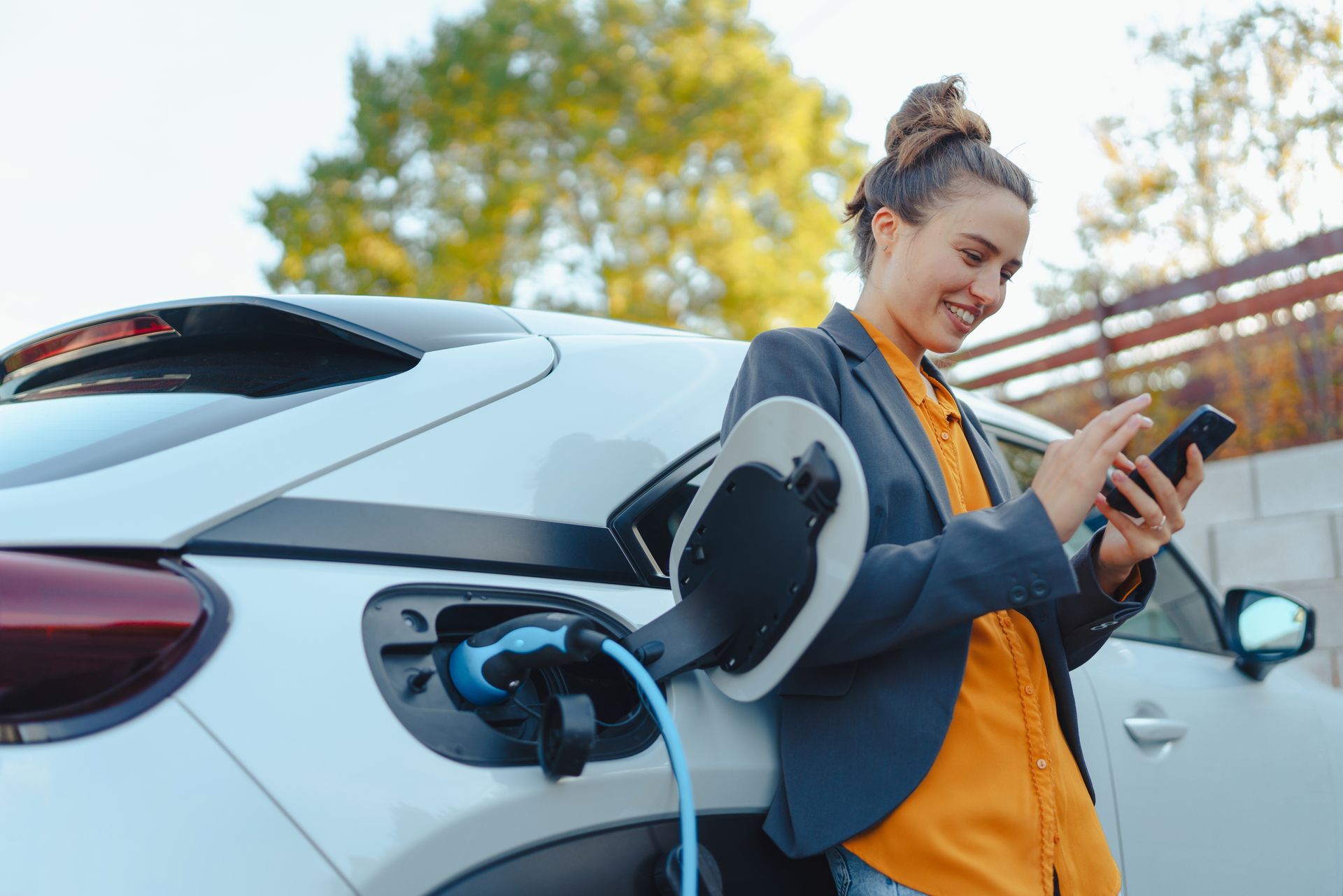 Femme souriante, utilisant son téléphone à côté d'une voiture électrique en charge. En extérieur, temps ensoleillé.