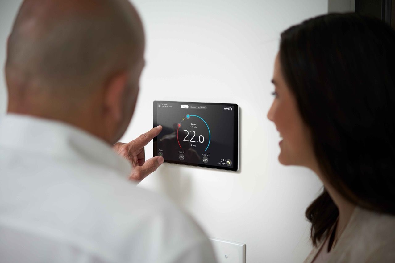 Un couple règle un thermostat intelligent fixé à un mur blanc, à l'intérieur. L'homme montre l'écran du doigt. La température est de 22 degrés.