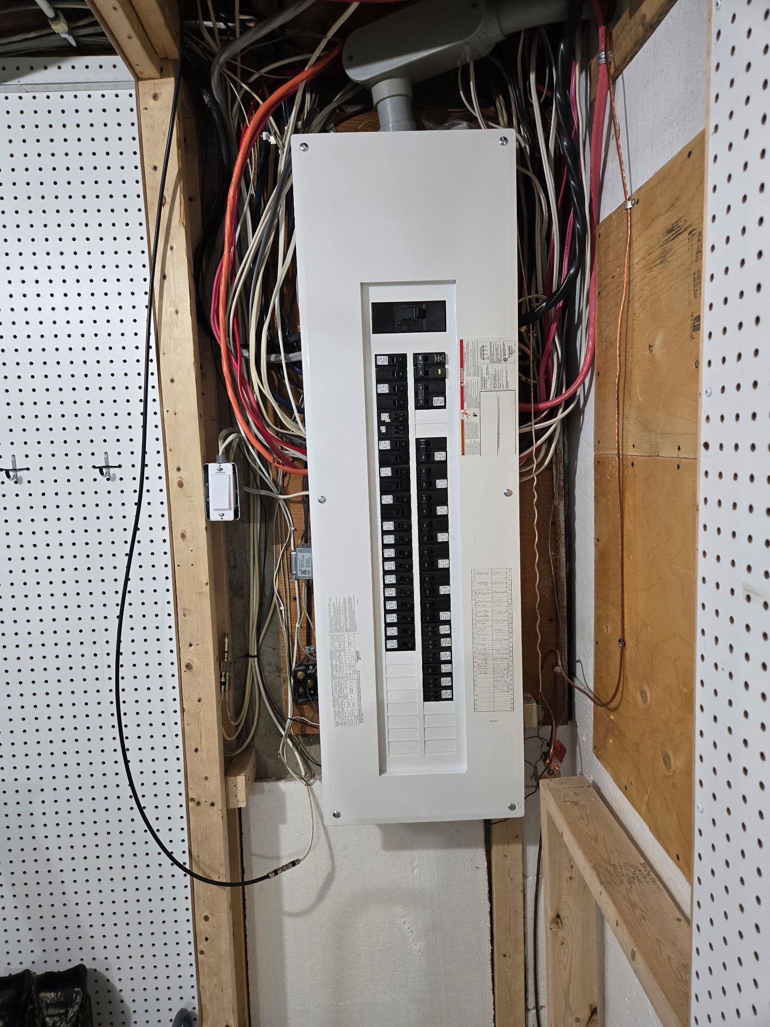 Un tableau électrique fixé au mur entre deux montants en bois, entouré de câbles apparents et de panneaux perforés.