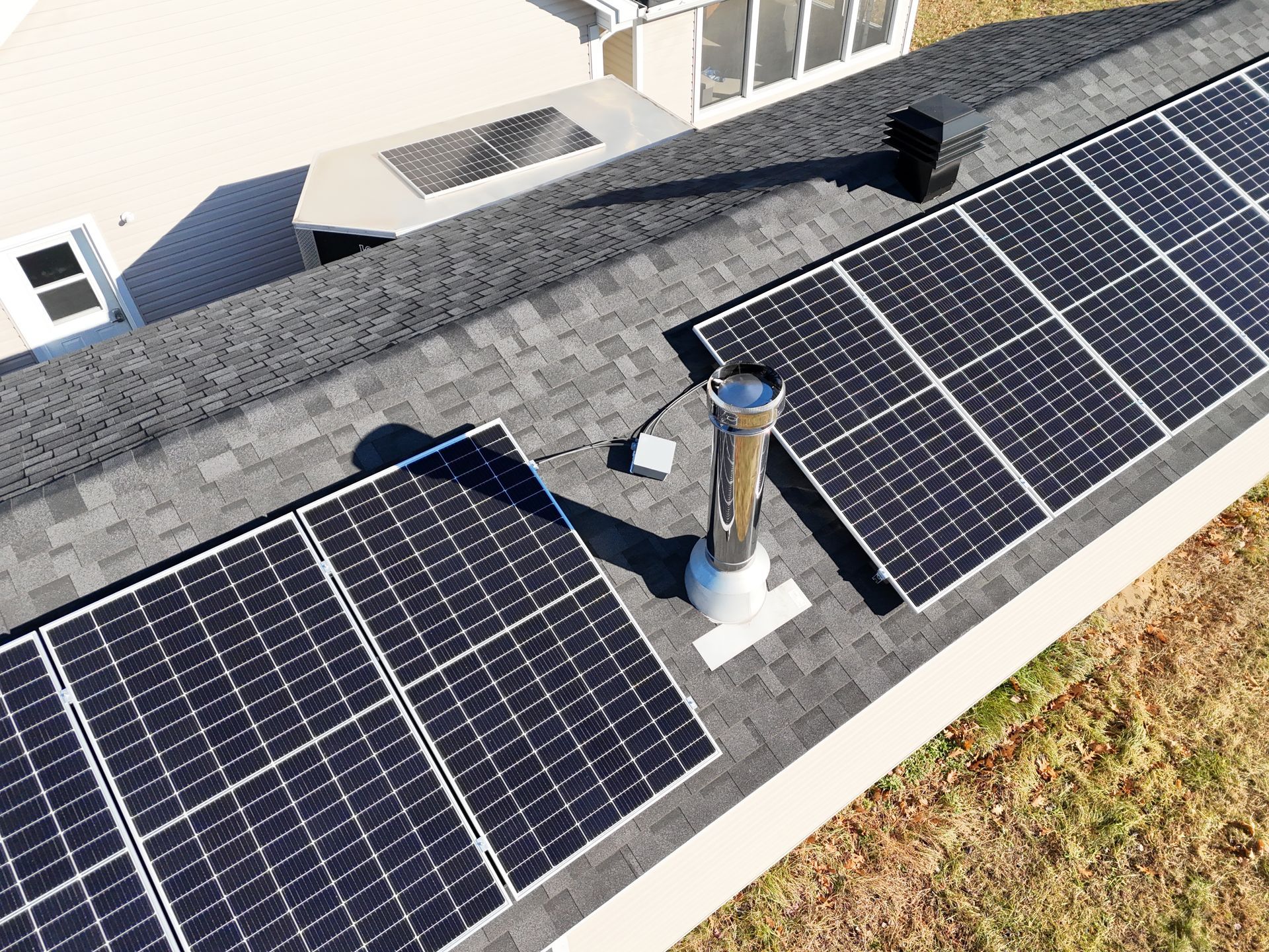 Vue de toit montrant des panneaux solaires sombres montés sur des bardeaux d'asphalte, avec un tuyau de ventilation métallique et des câbles au centre.