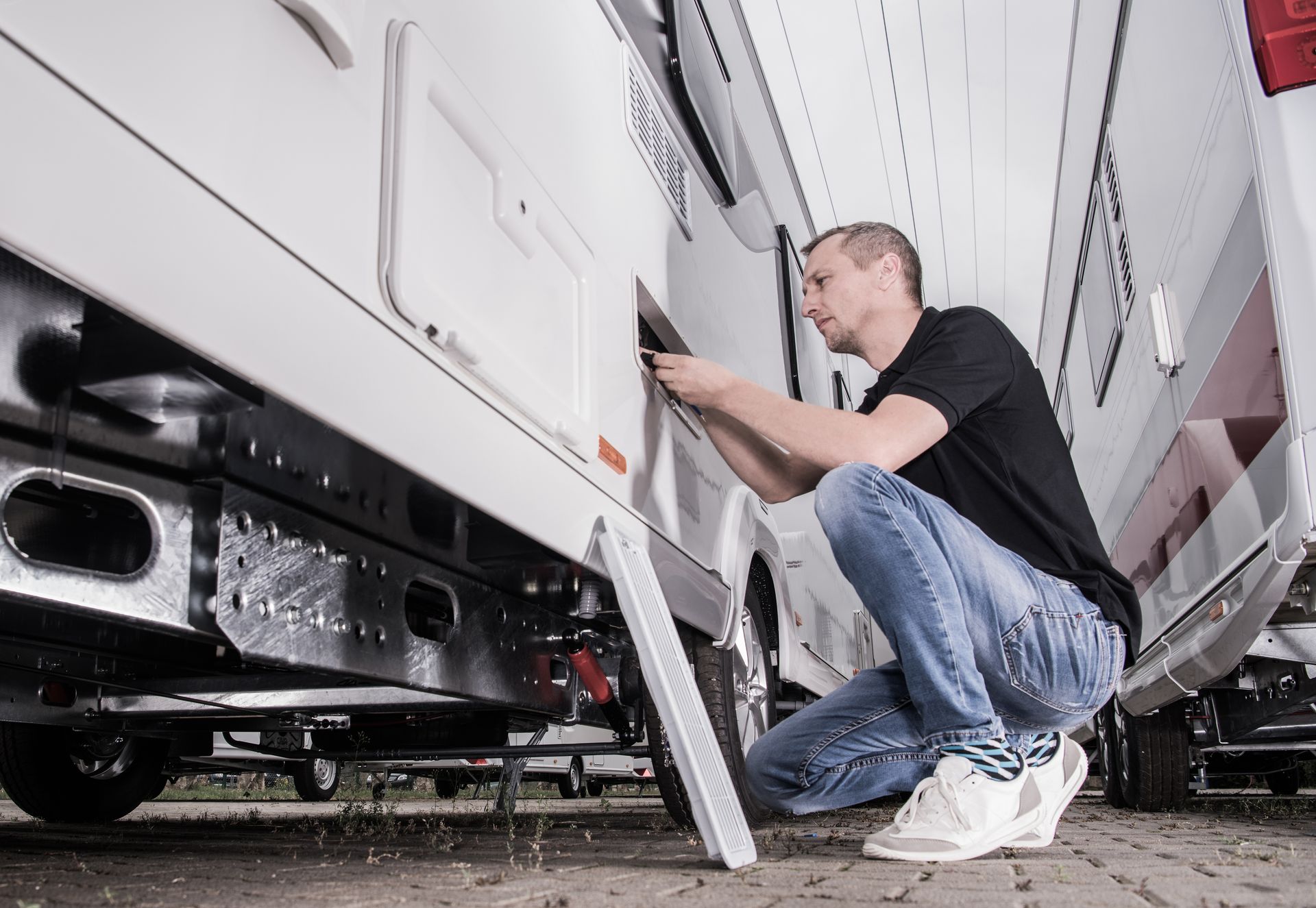 Man kneeling beside a white RV, inspecting or repairing the side panel with a tool