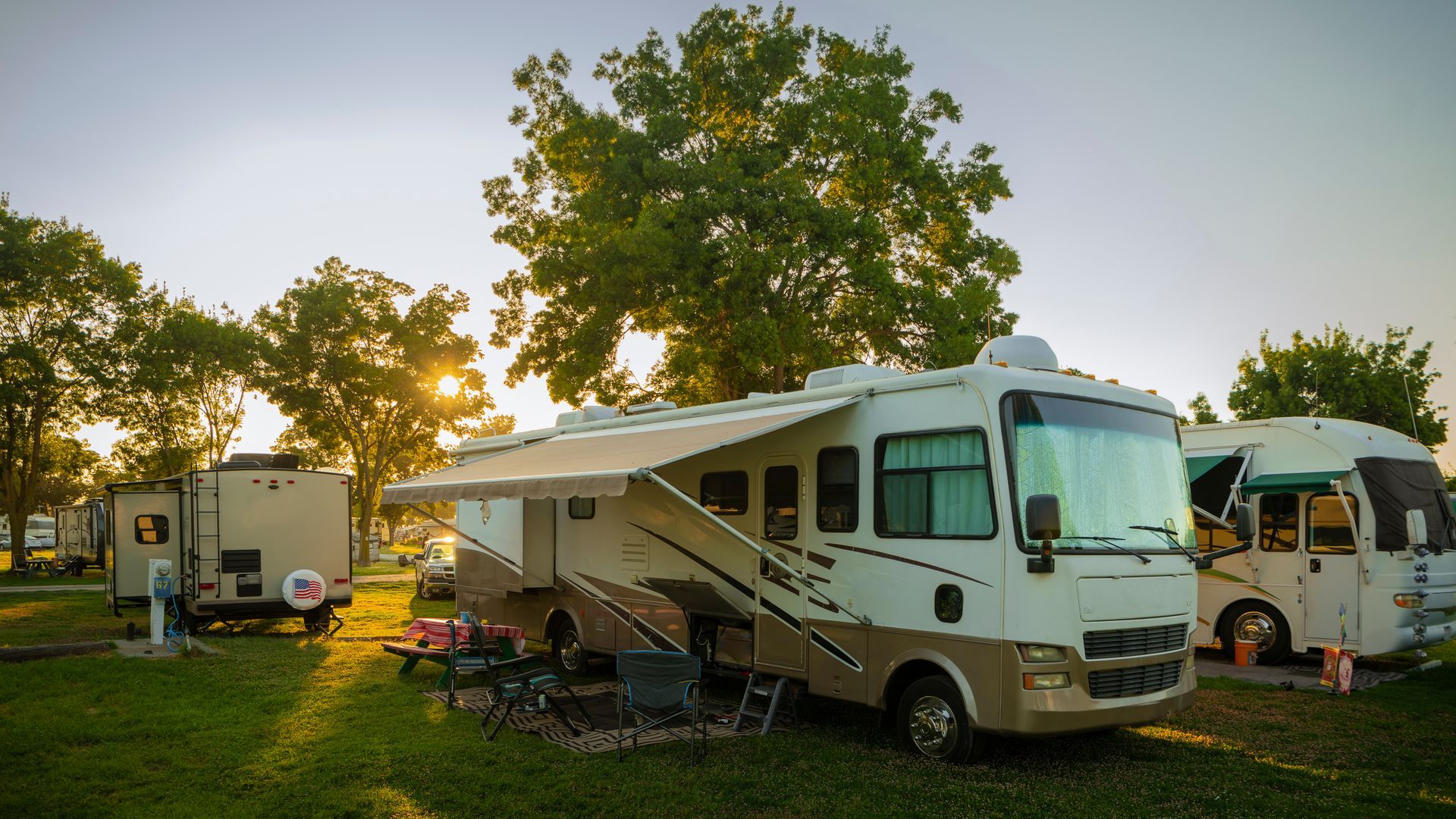 RV campers parked on grass at sunset under trees, with warm light and a white motorhome in front