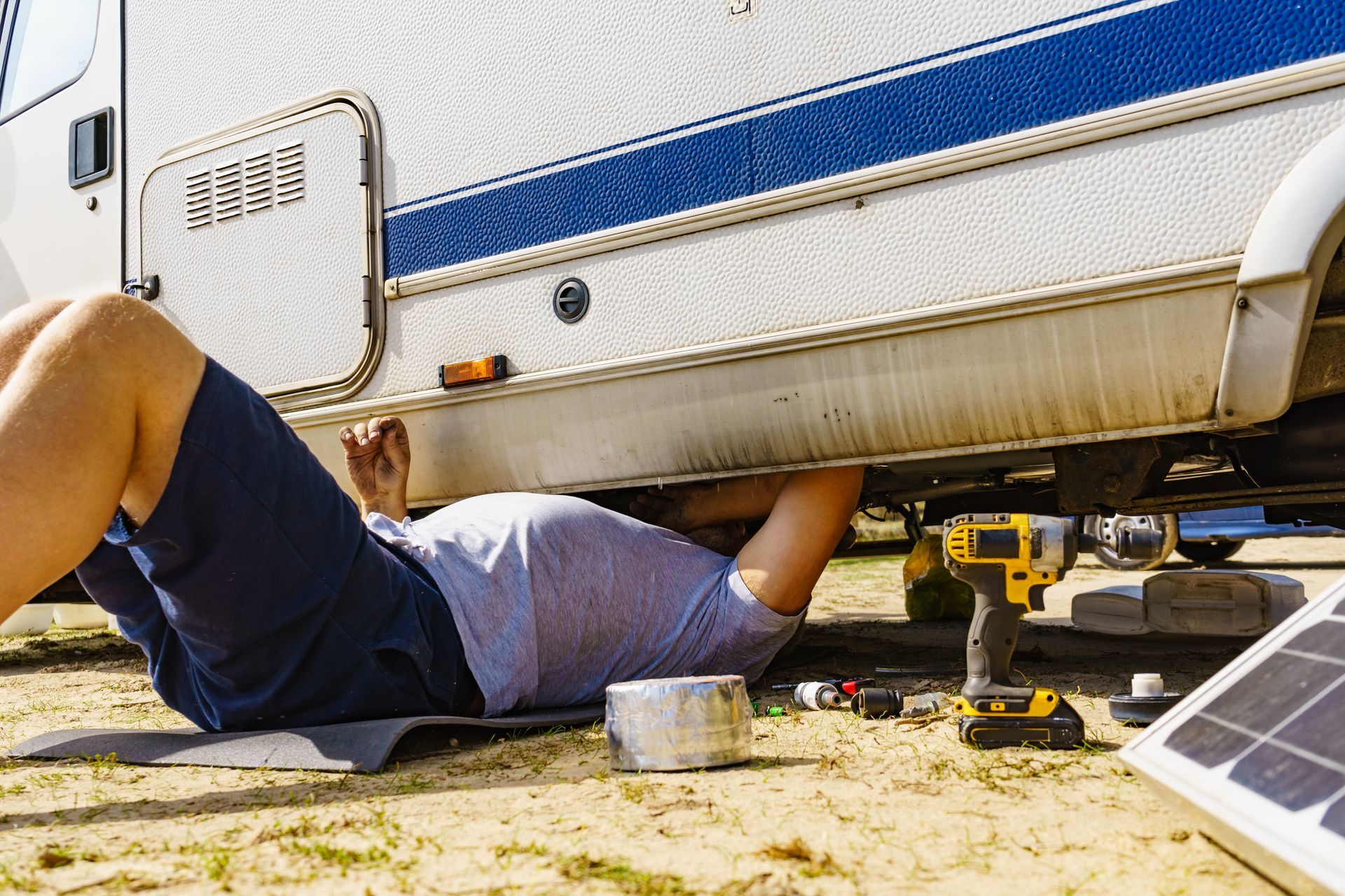 Person lying under a trailer while using tools near a solar panel on sandy ground