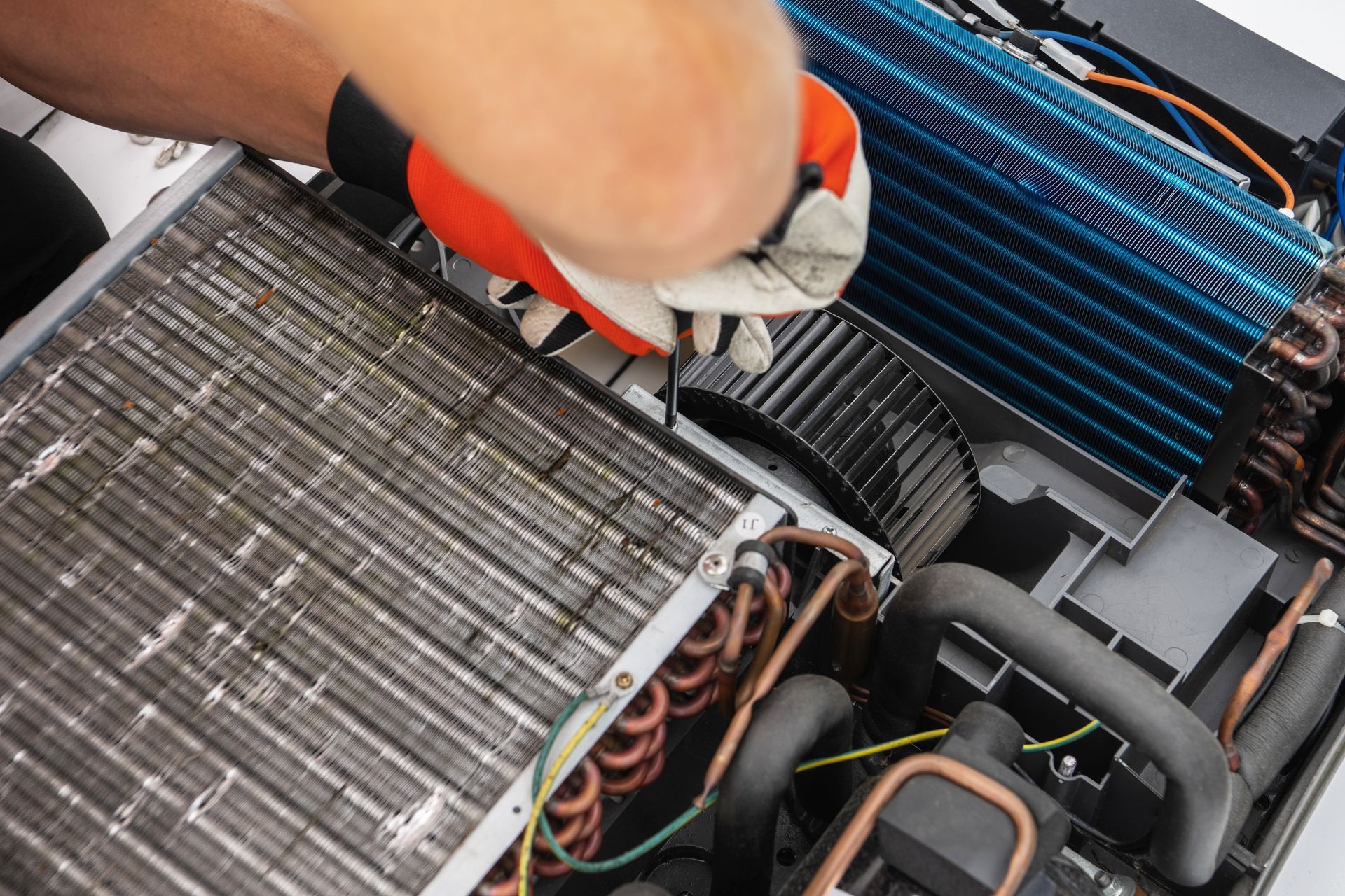 Gloved hand cleaning the fins of a dusty HVAC unit with a brush and hose.