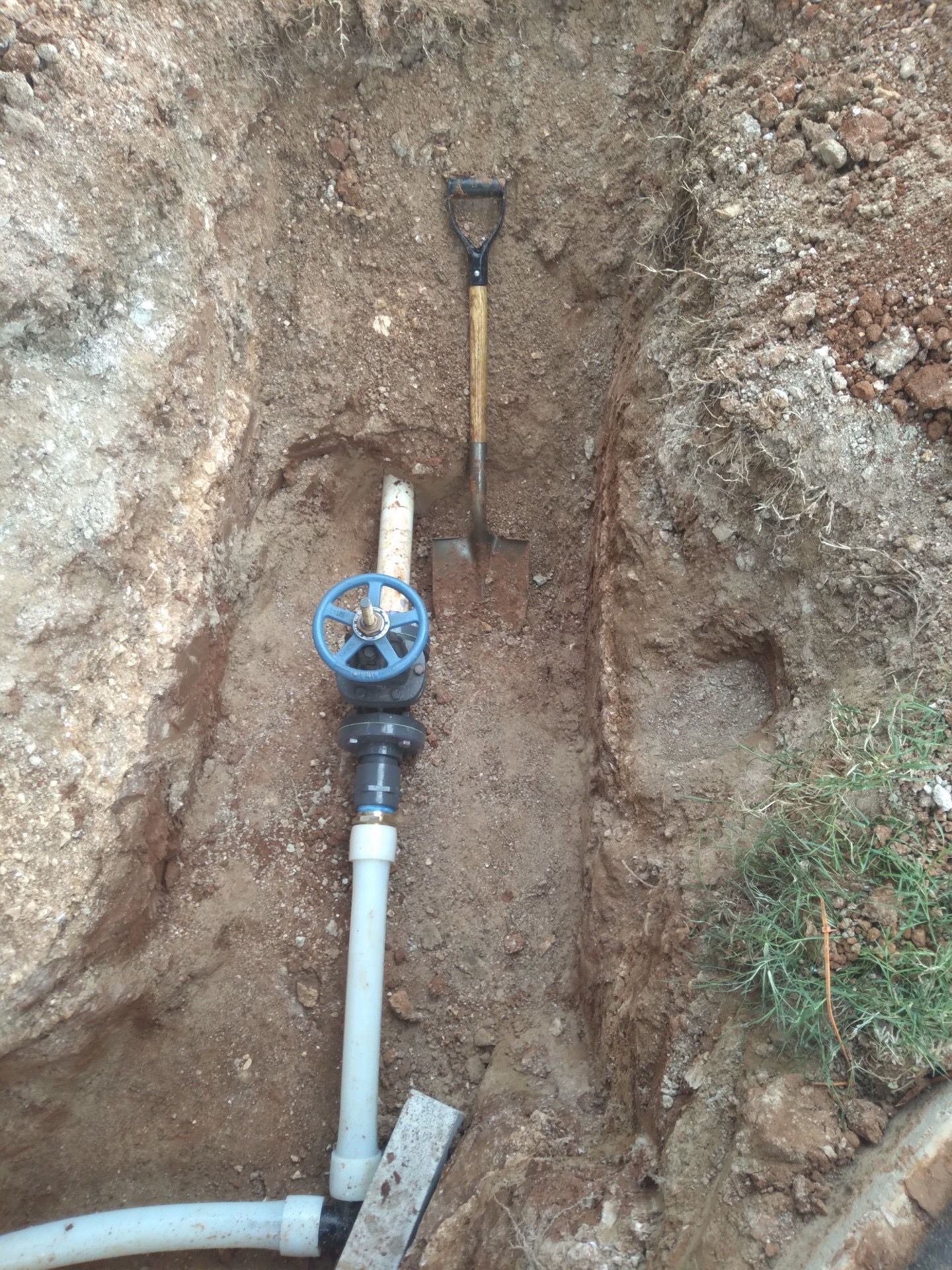 A buried sprinkler valve and piping exposed in a dug trench, with a shovel standing upright.