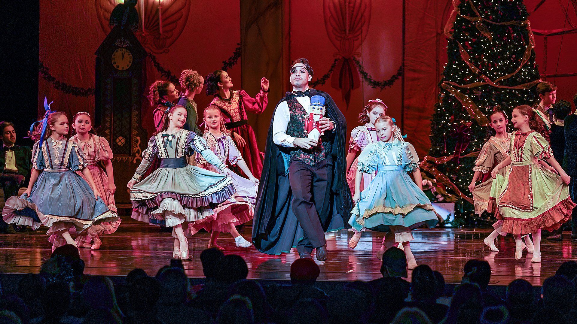 Drosselmeyer is holding the Nutcracker next to children in the Nutcracker Ballet by ACBallet.